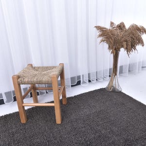 May include: A woven wicker stool with a wooden frame sits on a dark gray rug in front of a white sheer curtain. Dried pampas grass in a clear vase is on the right side of the image.