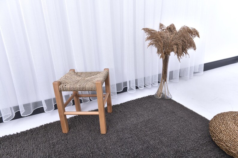 May include: A woven wicker stool with a wooden frame sits on a dark gray rug in front of a white sheer curtain. Dried pampas grass in a clear vase is on the right side of the image.