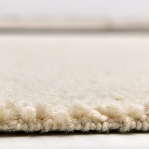 May include: Close-up of a cream-colored rug with a thick, textured pile. The rug's edge is visible, showing the dense fibers and construction. The neutral color and plush texture suggest comfort and warmth, suitable for home decor.
