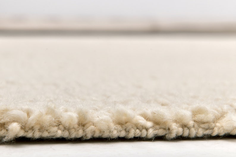May include: Close-up of a cream-colored rug with a thick, textured pile. The rug's edge is visible, showing the dense fibers and construction. The neutral color and plush texture suggest comfort and warmth, suitable for home decor.