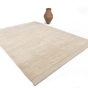 May include: A rectangular beige rug with a textured, grid-like pattern. A small, brown, terracotta vase sits upright on the white background. The rug's neutral color and design suggest a minimalist or natural aesthetic.
