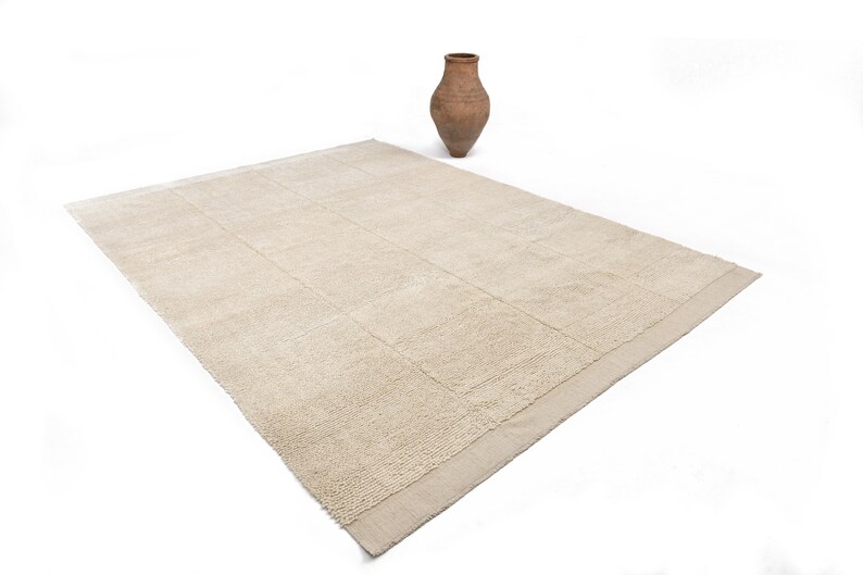 May include: A rectangular beige rug with a textured, grid-like pattern. A small, brown, terracotta vase sits upright on the white background. The rug's neutral color and design suggest a minimalist or natural aesthetic.