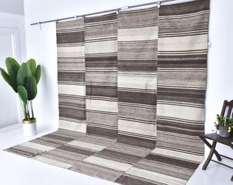 Op de afbeelding: Een groot, rechthoekig tapijt met horizontale strepen in tinten bruin, beige en wit. Het tapijt wordt opgehangen aan een metalen stang gepresenteerd, met een deel ervan op de vloer. Het ontwerp van het tapijt heeft een herhalend patroon van verschillende streepbreedtes.