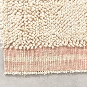 Puede incluir: Una alfombra de color crema con una textura esponjosa y un borde rayado rosa y crema.