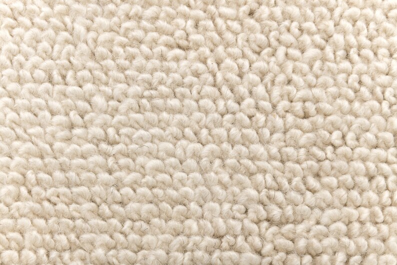 May include: Close-up of a cream-colored, textured fabric. The material has a dense, looped pile, creating a soft, plush appearance. The surface is covered in small, tightly packed loops, giving it a warm and inviting look. The fabric appears to be a type of faux fur or shearling.