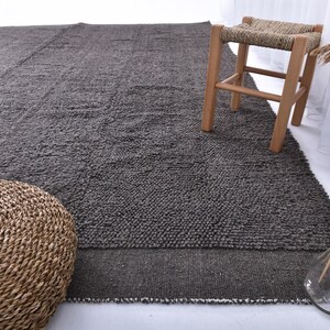 May include: A dark gray area rug with a textured surface. The rug is made of wool and has a natural, rustic look. A woven stool with a light wood frame is sitting on the rug.