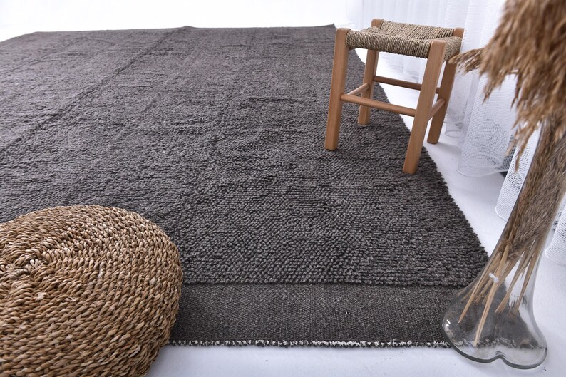May include: A dark gray area rug with a textured surface. The rug is made of wool and has a natural, rustic look. A woven stool with a light wood frame is sitting on the rug.