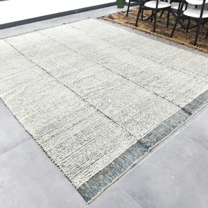 May include: A white and gray area rug with a blue border. The rug has a textured, woven look and is laid out on a gray tiled floor.