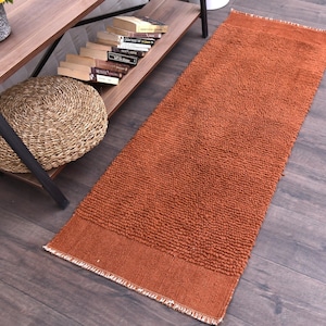 May include: An orange rectangular rug with a textured, looped pile. The rug has a solid border at the top and bottom with a fringed edge. The rug is placed on a dark wood floor.