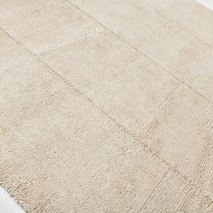 May include: A neutral-toned rug with a textured surface and a rectangular shape. The rug features a pattern of parallel lines, creating a subtle, geometric design. The color is a light beige, suitable for various interior styles.