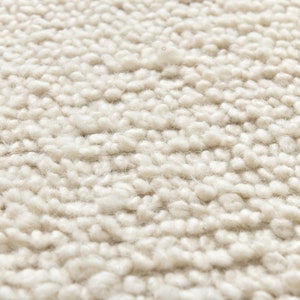 May include: Close-up of a cream-colored rug with a textured, fluffy surface. The rug is made of tightly woven loops of yarn, creating a soft and inviting texture.