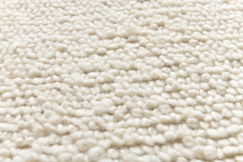 May include: Close-up of a cream-colored rug with a textured, fluffy surface. The rug is made of tightly woven loops of yarn, creating a soft and inviting texture.