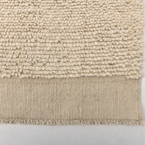 May include: Close-up of a beige rug with a textured, looped pile. The lower section features a woven, flat-weave border with a fringed edge. The rug's neutral color and texture suggest a cozy, natural aesthetic.