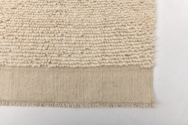May include: Close-up of a beige rug with a textured, looped pile. The lower section features a woven, flat-weave border with a fringed edge. The rug's neutral color and texture suggest a cozy, natural aesthetic.