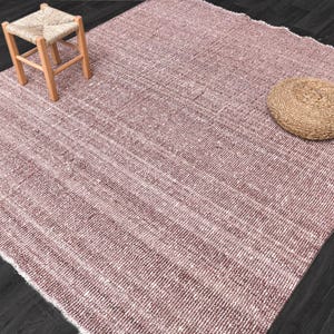 Op de afbeelding: Een roze en wit geweven tapijt met een natuurlijke geweven kruk en een rond geweven kussen op het tapijt.
