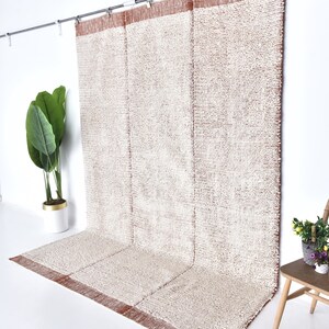 May include: A white and brown woven rug with a textured surface. The rug is hanging on a rod and has a brown border.