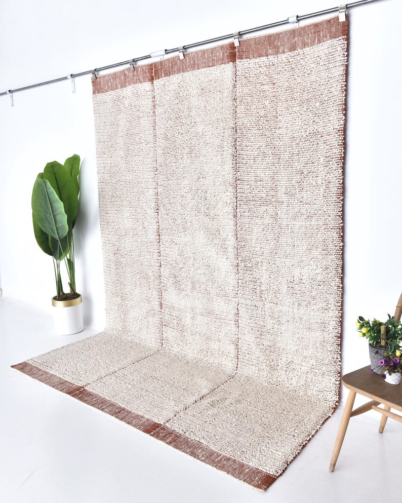 May include: A white and brown woven rug with a textured surface. The rug is hanging on a rod and has a brown border.