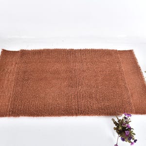 Puede incluir: Una alfombra de baño rectangular marrón con una superficie texturizada. La alfombra está hecha de un material suave y tiene un borde con flecos en ambos extremos. La alfombra está colocada sobre una superficie blanca.