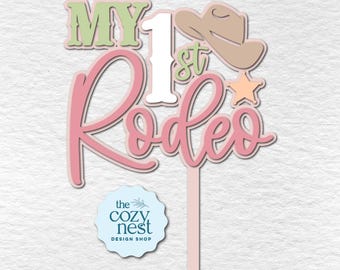 May include: A layered cake topper with the words "My 1st Rodeo" in pink and green, a cowboy hat, and a star. The topper is designed for a first birthday celebration. The Cozy Nest Design Shop logo is at the bottom.