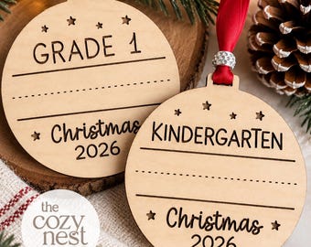 Puede incluir: Dos adornos redondos de madera con detalles de estrellas cortadas a láser y cintas. Uno está grabado con GRADE 1 y el otro con KINDERGARTEN. Ambos incluyen líneas para escribir y el texto Christmas 2026, con cintas de color rosa y rojo.