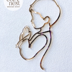 Può includere: Un disegno tagliato al laser color oro di una persona in preghiera, con un rosario. Il disegno è una silhouette, con la testa e le mani di profilo. Il testo "SVG LASER FILE" è in basso.