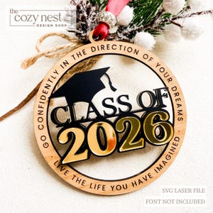 Könnte beinhalten: Runder Holz-Abschlussornament mit der Aufschrift "Class of 2026" in Gold und Schwarz. Der Rand des Ornaments trägt die Inschriften "Go confidently in the direction of your dreams" und "Live the life you have imagined".