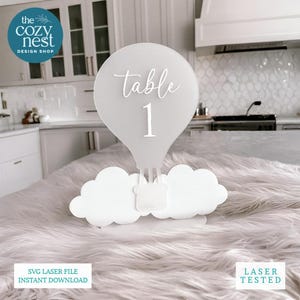 May include: A white laser-cut hot air balloon table number with the text "table 1". The balloon sits atop two cloud-shaped bases. The Cozy Nest Design Shop logo is in the upper left corner. The bottom of the image has the text "SVG Laser File Instant Download" and "Laser Tested".