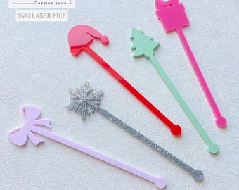 Op de afbeelding: Een verzameling kleurrijke acryl roerstaafjes in feestelijke vormen: een kerstmuts, kerstboom, cadeauverpakking, strik en sneeuwvlok. De roerstaafjes zijn in tinten roze, rood, groen en zilver glitter. De tekst "the cozy nest design shop" en "SVG laser file" zijn zichtbaar.