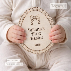 Könnte beinhalten: Ein hölzernes Osterei-Schild mit einem gewellten Rand und einem Schleifendesign. Der Text "Juliana's First Easter 2026" ist auf dem Schild eingraviert. Das Schild wird von einem Baby gehalten, das einen weißen Strampler trägt. Das Cozy Nest Design Shop-Logo befindet sich oben rechts.