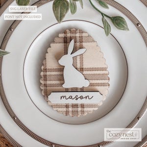 May include: A wooden Easter egg-shaped place card with a plaid pattern, topped with a white bunny silhouette. A small rectangular name tag with the name "mason" is below the bunny. The plate is white with a decorative rim.