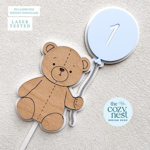 Könnte beinhalten: Ein Tortenaufsatz aus Holz mit einem Teddybären, der einen hellblauen Ballon mit der ausgeschnittenen Zahl 1 hält. Der Bär ist hellbraun mit weißer Kontur. Der Text "SVG LASER FILE INSTANT DOWNLOAD" und "LASER TESTED" befindet sich oben links.