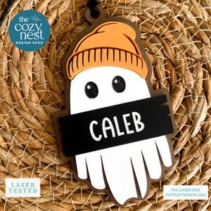 May include: A wooden ghost-shaped ornament with an orange beanie and the name "CALEB" on a black banner. The Cozy Nest Design Shop logo is in the upper left corner. The ornament is laser tested and an SVG laser file is available.