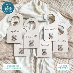 以下が含まれることがあります： 木製のベビー服ハンガー。彫刻された文字とテディベアのデザインが特徴です。ハンガーには、新生児、0〜3か月、3〜6か月、6〜9か月、9〜12か月、12〜18か月の年齢範囲が記載されています。The Cozy Nest Design Shop.