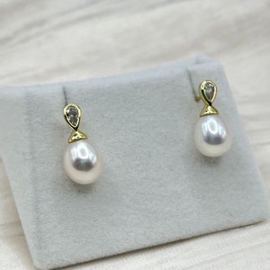 May include: A pair of teardrop-shaped pearl earrings. Each earring features a lustrous white pearl suspended from a gold-colored setting with a small, clear gemstone accent. The earrings are displayed on a light gray surface.