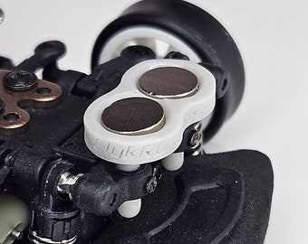 Puede incluir: Primer plano de un chasis de coche de carreras en miniatura en blanco y negro con un parachoques delantero blanco y dos imanes negros unidos a la parte delantera.