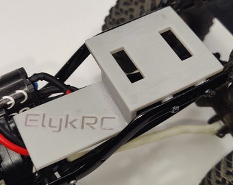 May include: A black and white RC car chassis with a gray 3D printed battery tray. The tray has two slots for batteries and the text "ElyK RC" printed on it. The car has black wheels with a tread pattern and white wheels on the suspension.