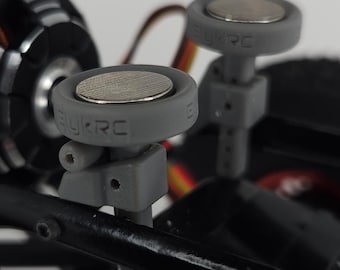 May include: A radio-controlled vehicle chassis featuring a black and red electric motor, black suspension components with red accents, and two grey circular body mounts with magnetic tops, each embossed with the text BYRC.