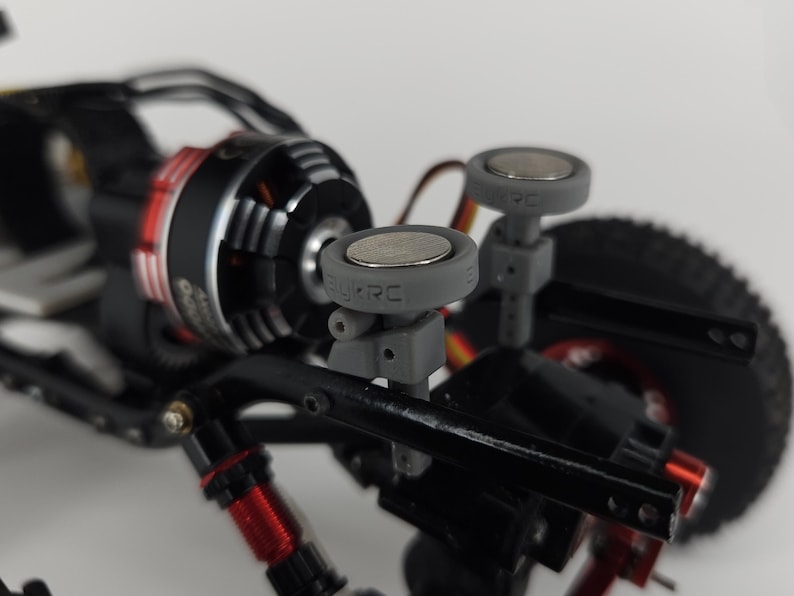 May include: A radio-controlled vehicle chassis featuring a black and red electric motor, black suspension components with red accents, and two grey circular body mounts with magnetic tops, each embossed with the text BYRC.
