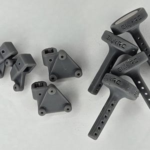 May include: A collection of dark gray 3D printed components for remote control vehicles, including four triangular mounting brackets with circular holes and three T-shaped antenna mounts featuring the embossed text ElykRC.