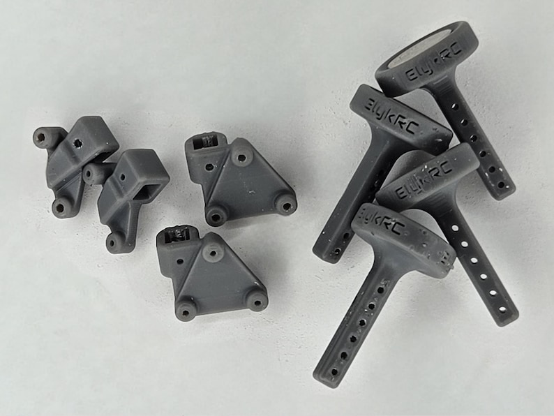 May include: A collection of dark gray 3D printed components for remote control vehicles, including four triangular mounting brackets with circular holes and three T-shaped antenna mounts featuring the embossed text ElykRC.
