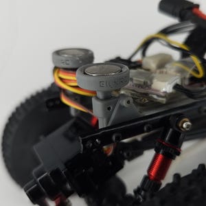May include: A remote-controlled vehicle chassis featuring two grey circular mounts with the text EILINRC embossed on the side. The assembly includes black structural components, red metallic shock absorbers, black knobby tires, and exposed wiring.