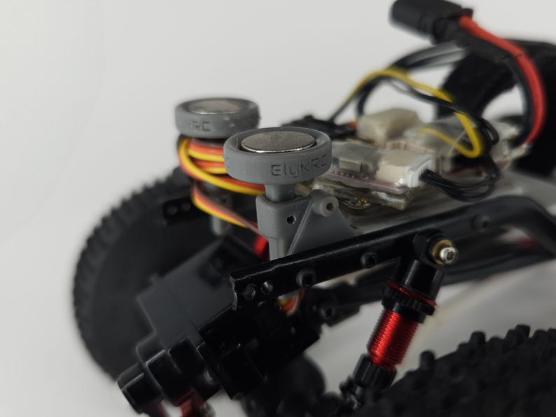 May include: A remote-controlled vehicle chassis featuring two grey circular mounts with the text EILINRC embossed on the side. The assembly includes black structural components, red metallic shock absorbers, black knobby tires, and exposed wiring.