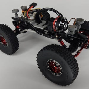 May include: A remote-controlled rock crawler chassis featuring a black metal frame, red anodized aluminum wheel hubs, and large knobby black rubber tires. The vehicle includes a brushless electric motor, exposed wiring, and four grey circular body mounts.