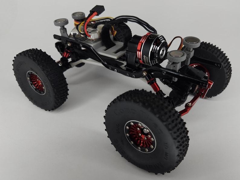 May include: A remote-controlled rock crawler chassis featuring a black metal frame, red anodized aluminum wheel hubs, and large knobby black rubber tires. The vehicle includes a brushless electric motor, exposed wiring, and four grey circular body mounts.
