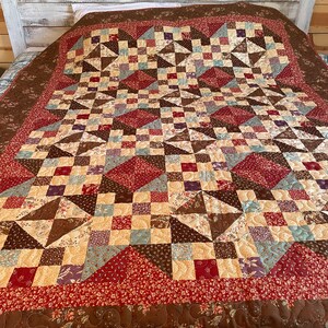May include: A brown, red and cream patchwork quilt with a geometric pattern. The quilt is made of various fabrics with floral prints and is displayed on a bed.