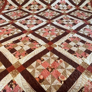 May include: A quilt with a brown, cream, and pink floral pattern. The quilt is made up of squares with a diamond pattern in the center. The quilt is quilted with a decorative stitch.