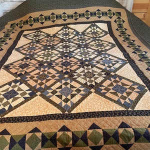 May include: A large, handmade quilt with a geometric pattern in shades of beige, green, and navy blue. The quilt features diamond and square shapes, with a dark green border. The quilt is displayed on a bed.