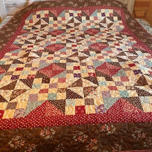 May include: A brown, red and blue patchwork quilt with a floral pattern on the border. The quilt is on a bed with a white headboard and a plaid bed skirt.