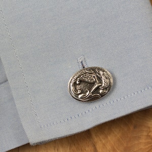 May include: Silver cufflinks with a detailed design of a mythical creature, possibly a dragon or griffin, with wings and a curled tail.