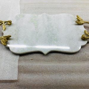 May include: A rectangular marble serving tray with a scalloped edge and gold-coloured branch handles. The tray is white with subtle green veining, and the handles are detailed with leaves and stems. A decorative piece for serving.